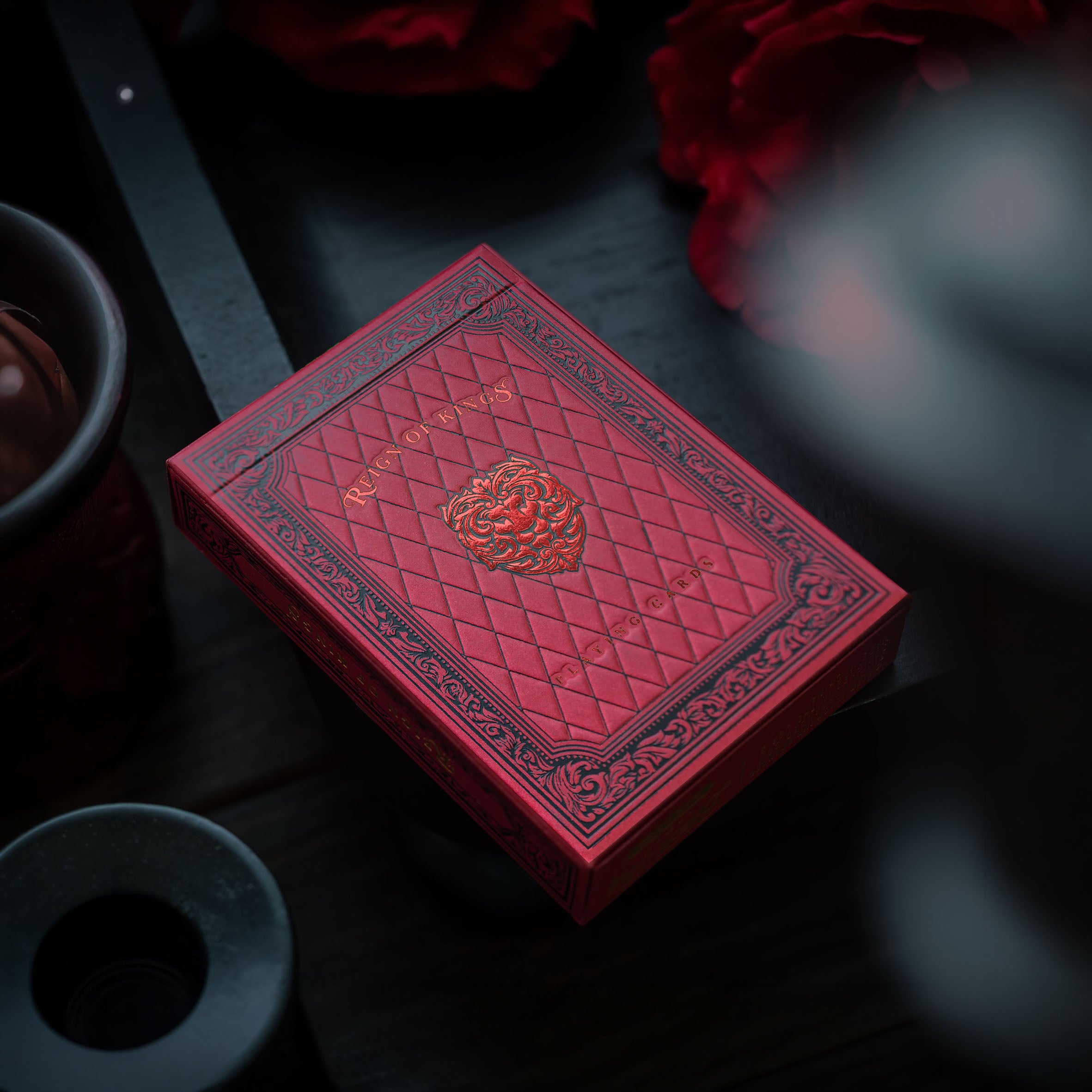 A red playing card deck box with embossed diamond quilted texture and ornate border detailing, photographed on a dark tabletop with a blurred red flower in the background.