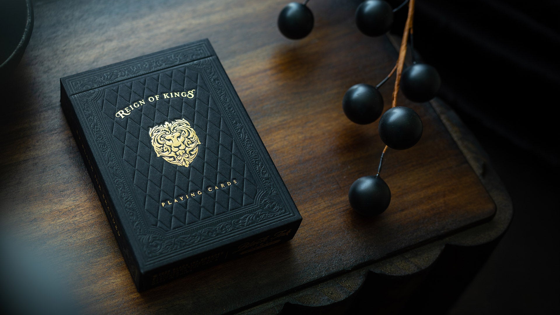 A black embossed deck for 'Reign of Kings Playing Cards' with gold lettering and crest, resting on a dark wooden surface next to a decorative black beaded garland.